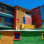 Casas coloridas de madeira em um bairro tradicional de Buenos Aires, com fachadas pintadas em azul, verde, vermelho e amarelo sob um c&eacute;u azul.