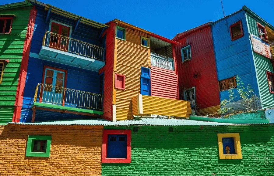 Casas coloridas de madeira em um bairro tradicional de Buenos Aires, com fachadas pintadas em azul, verde, vermelho e amarelo sob um céu azul.