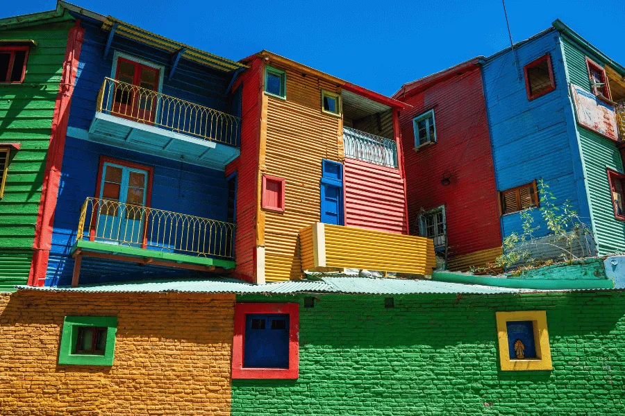 Casas coloridas de madeira em um bairro tradicional de Buenos Aires, com fachadas pintadas em azul, verde, vermelho e amarelo sob um céu azul.